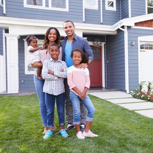 Happy family of five—mom, dad, and three young kids—standing on green grass in front of a blue house in the Chandler Creek neighborhood near Austin, TX.